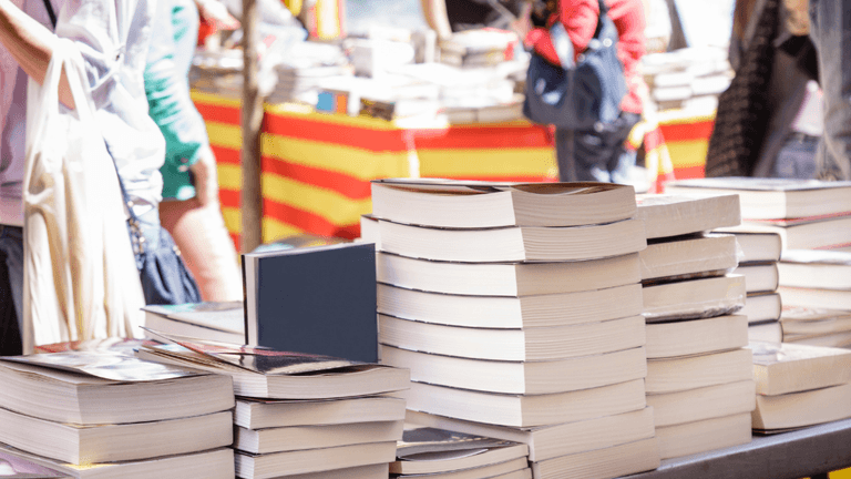Stacks of books on a table with blurred vendors and colorful backgrounds in a market